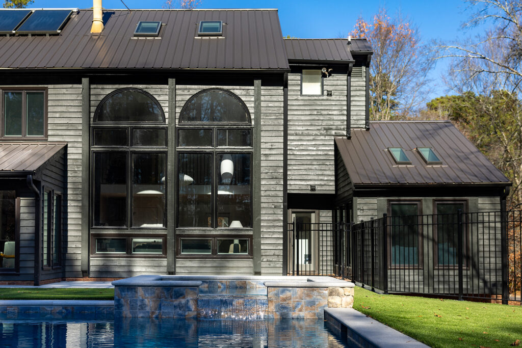 Exterior of the back of a renovated two-story gray home with two two-story tall round-top picture windows and an inground pool in the foreground.